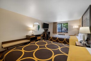 A hotel room with two beds, a desk, a chair, a television, and a window with a view of trees.