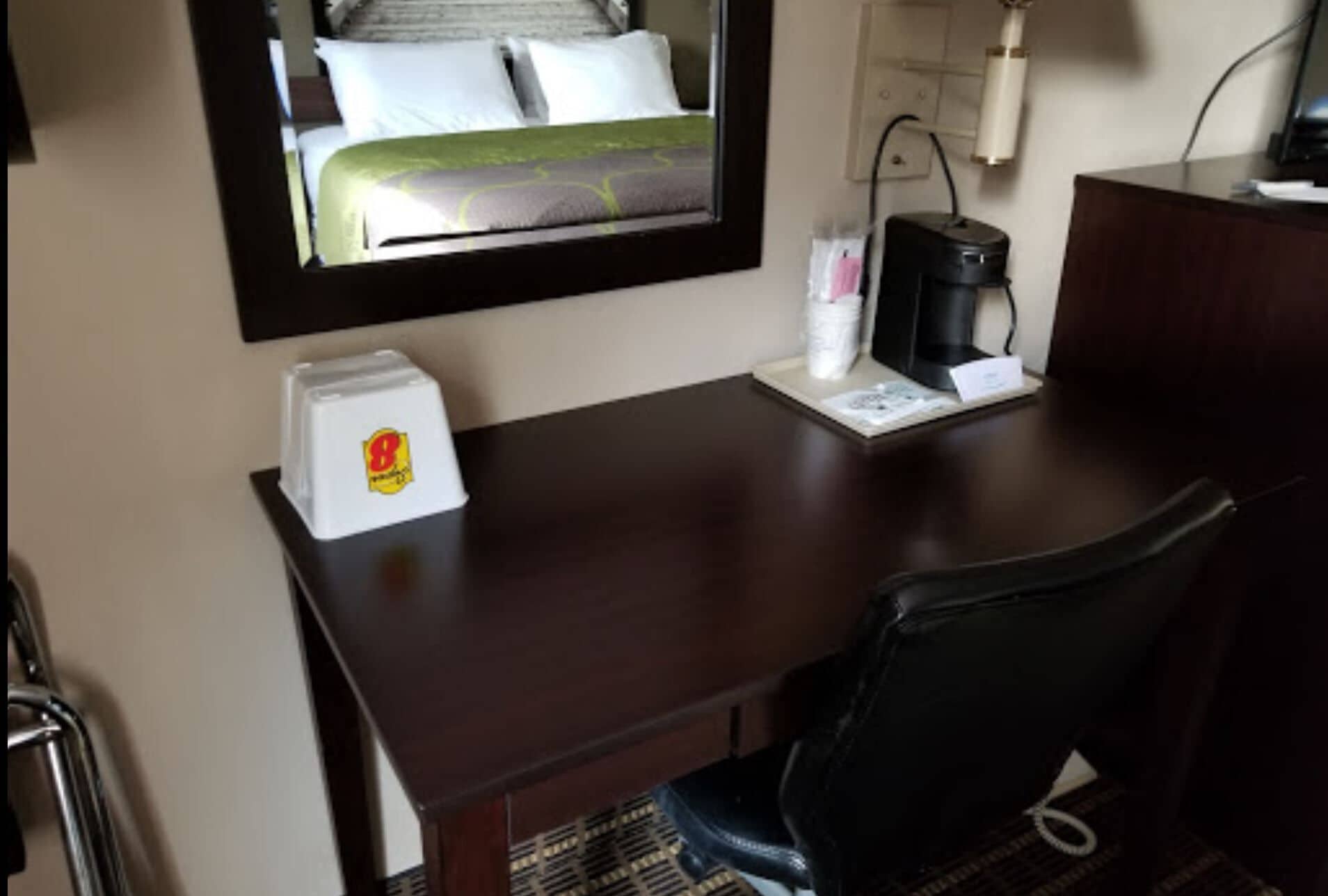A hotel room with a bed, a desk with a coffee maker, a chair, and a small table with a bottle and a menu.