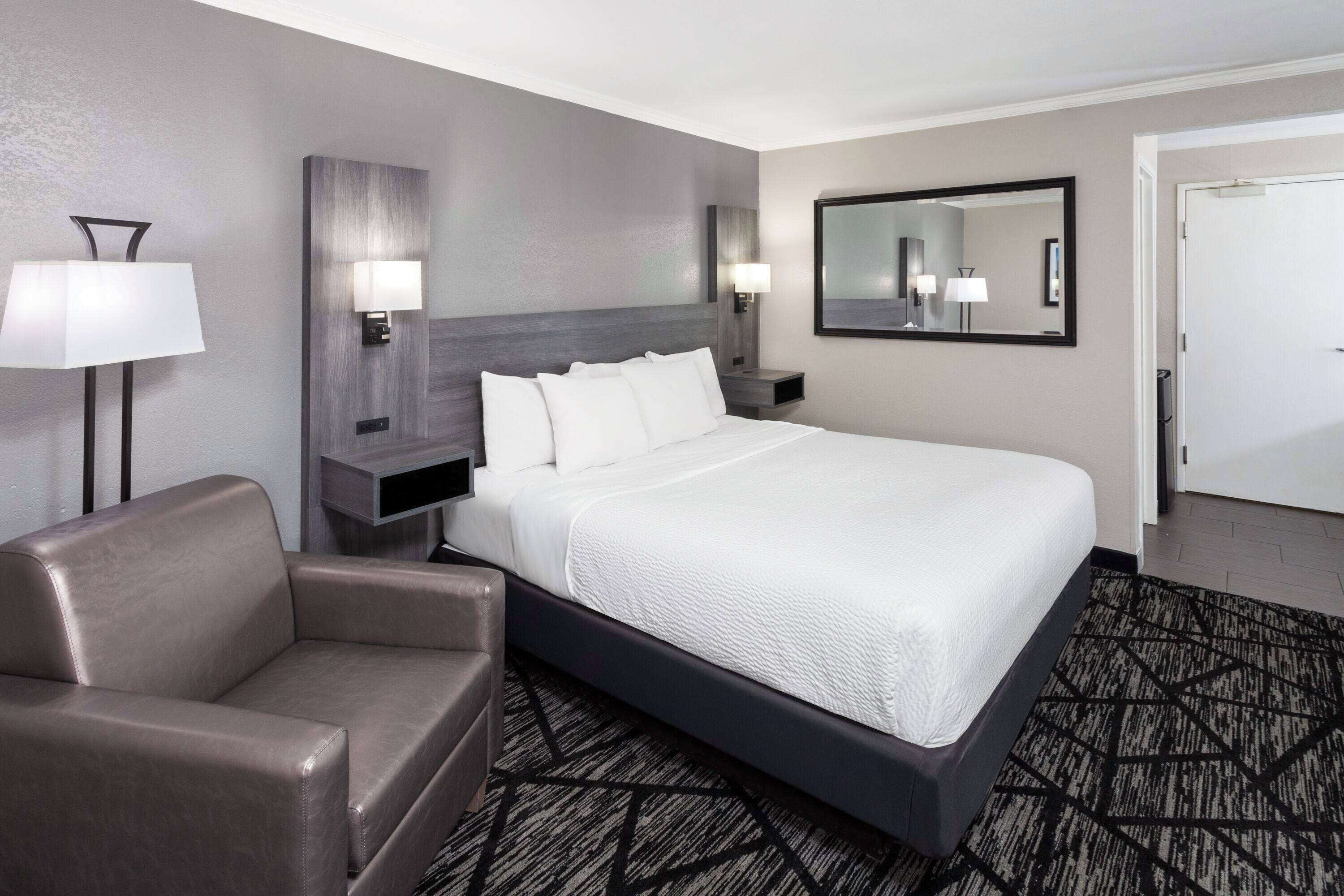 A modern hotel room with a large bed, a sofa, a nightstand, a wall-mounted lamp, and a mirror.