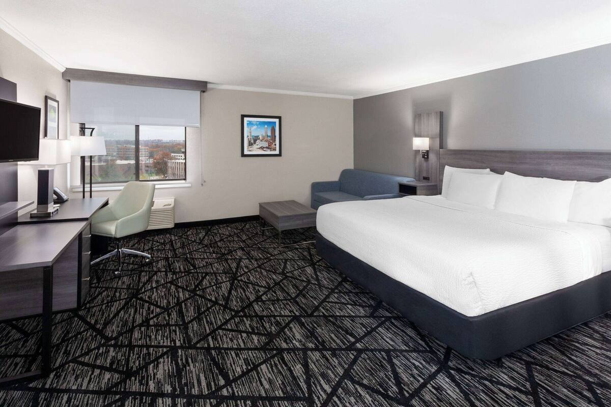 A hotel room with a large bed, a desk with a computer, a chair, and a window with a city view.