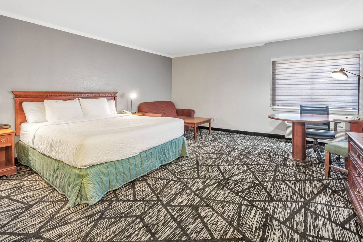 A hotel room with a bed, a desk, a chair, a lamp, and a window with blinds.