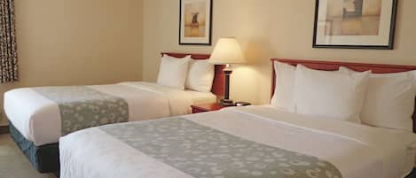 A hotel room with two beds, each with a pillow and a blanket. There is a nightstand with a lamp and a phone. The room has a carpeted floor and beige walls.