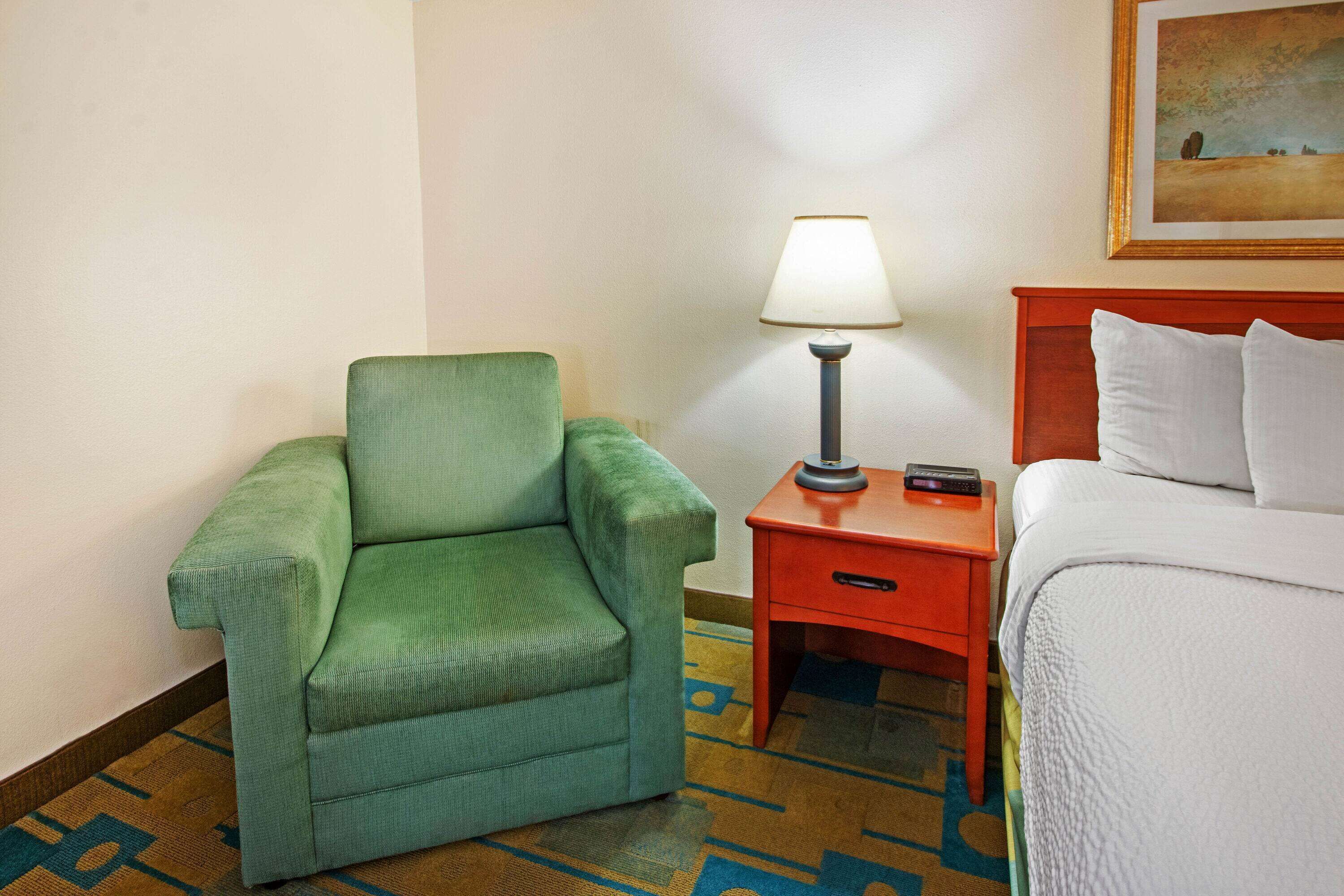 Une chambre d’hôtel avec un fauteuil vert, une petite table de chevet en bois avec une lampe et un lit avec une literie blanche.
