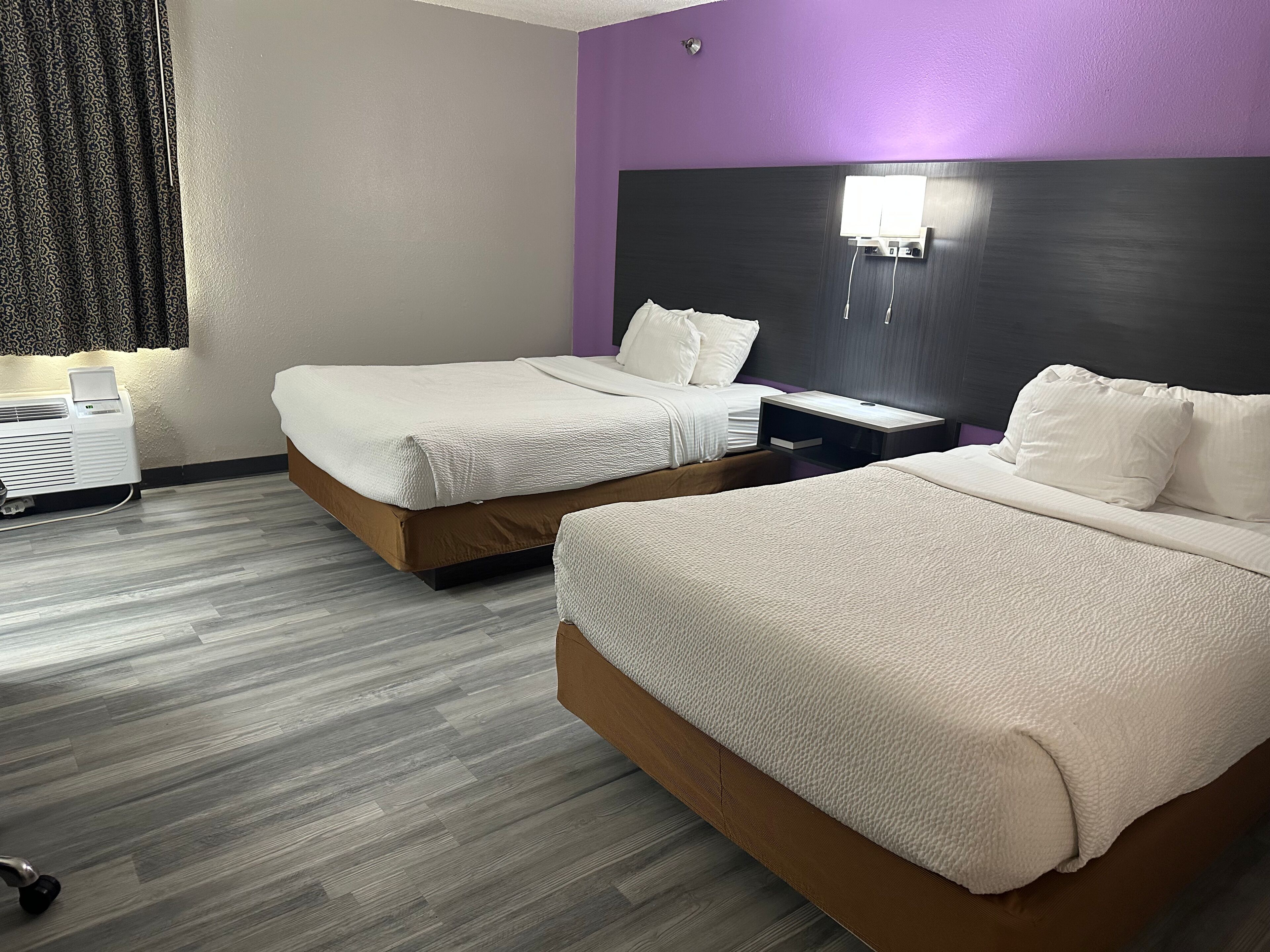 Habitació d'hotel amb dos llits, una taula de nit, una làmpada penjada a la paret i una finestra amb cortines.