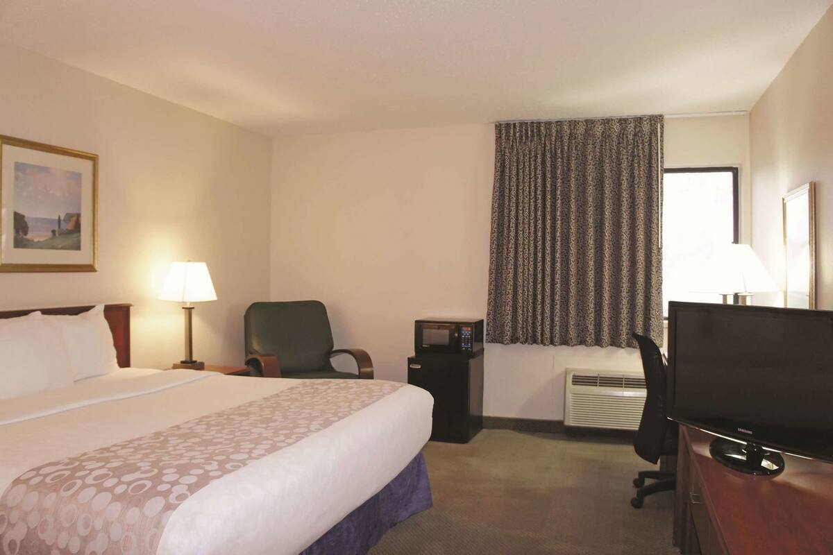A hotel room with a bed, a desk, a chair, a television, a lamp, and a window with curtains.