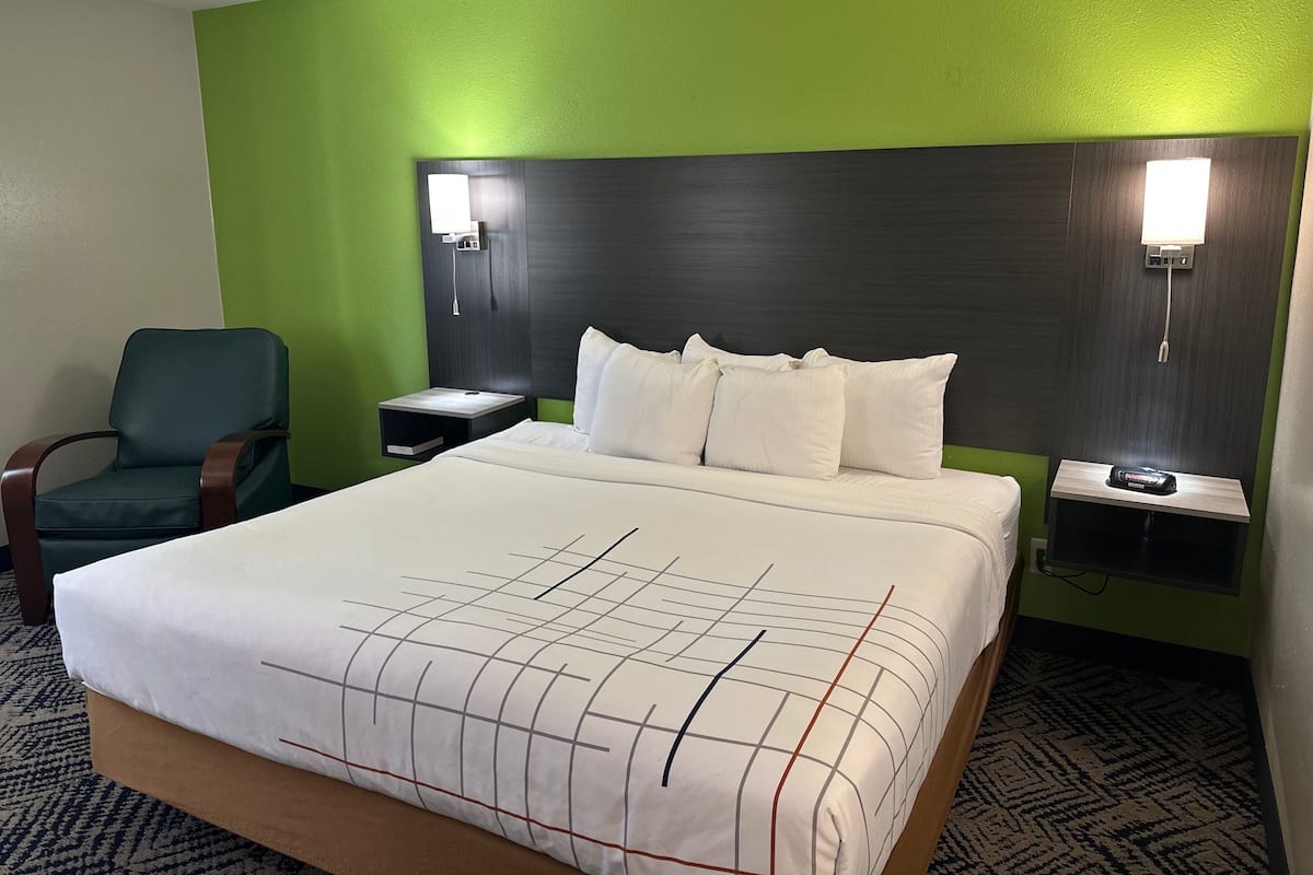 A hotel room with a large bed, green walls, a wooden headboard, two bedside tables with lamps, a chair, and a patterned carpet.