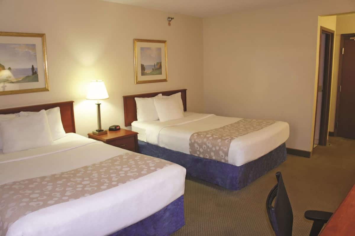 A hotel room with two beds, a nightstand with a lamp, a chair, and two framed pictures on the wall.