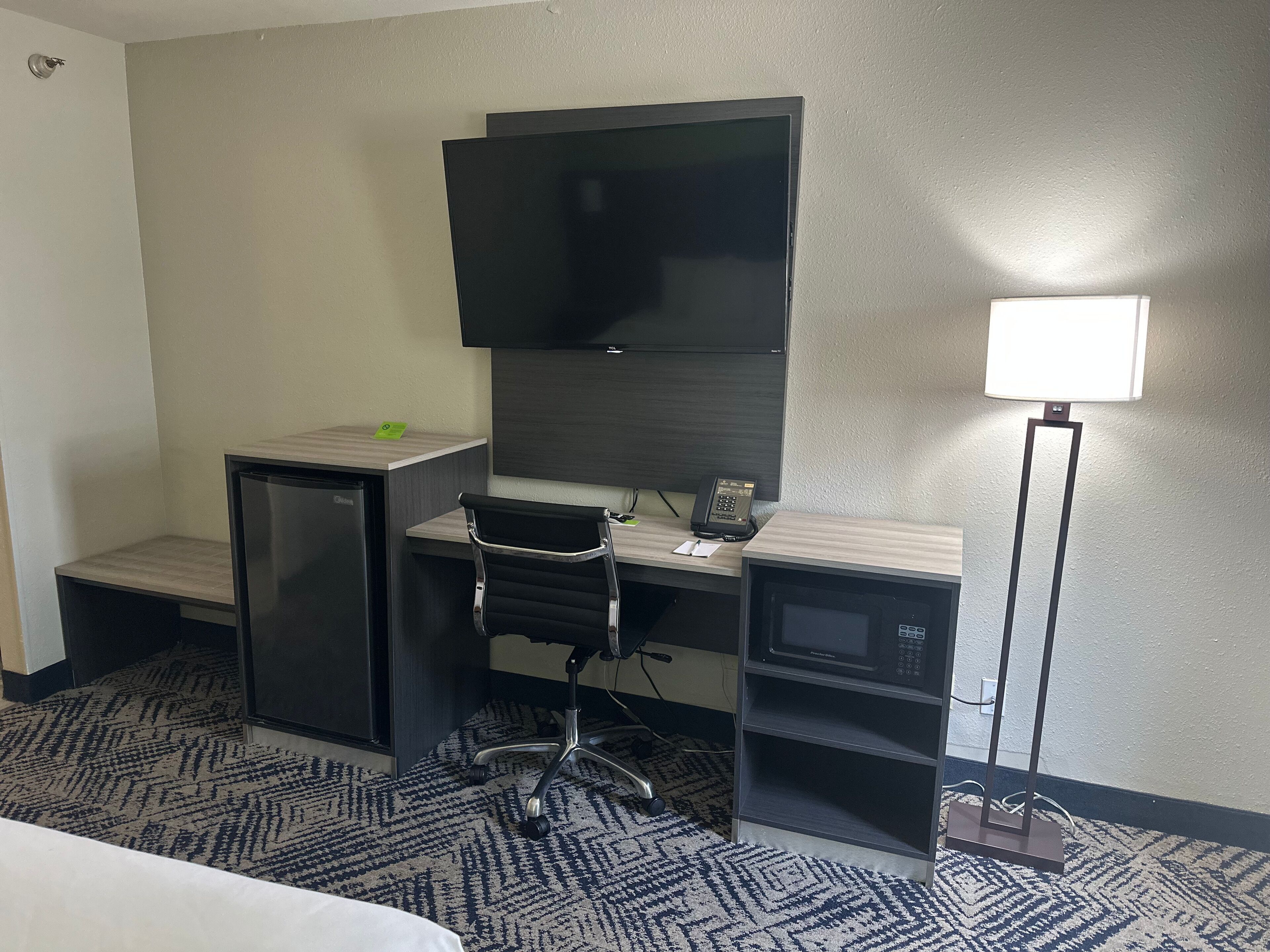 Habitación de hotel con escritorio, televisor de pantalla plana, refrigerador pequeño, microondas, lámpara de escritorio y alfombra estampada.