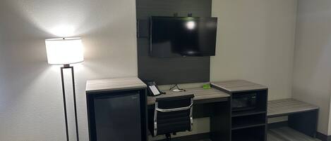 A hotel room with a desk, chair, TV, mini-fridge, lamp, and a bed.