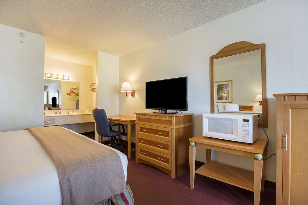 A hotel room with a bed, a desk with a computer, a television, a microwave, a mirror, and a dresser.