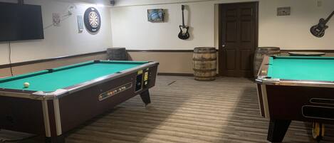 A pool table with a dartboard and a guitar, a wooden barrel, and a door in a room with carpeted flooring and overhead lighting.