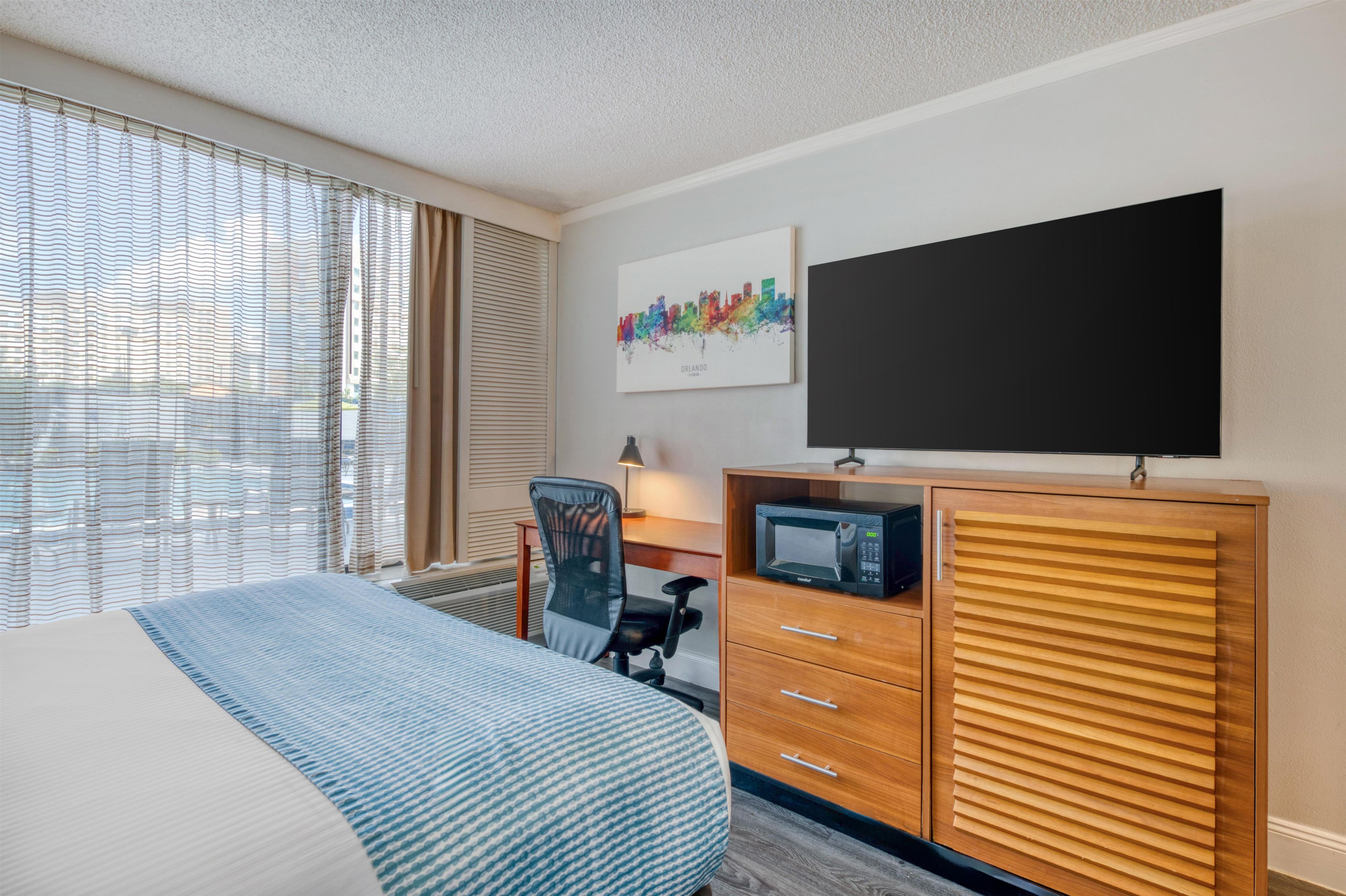 A hotel room with a bed, a desk with a computer, a chair, a flat-screen TV, and a wall-mounted artwork.