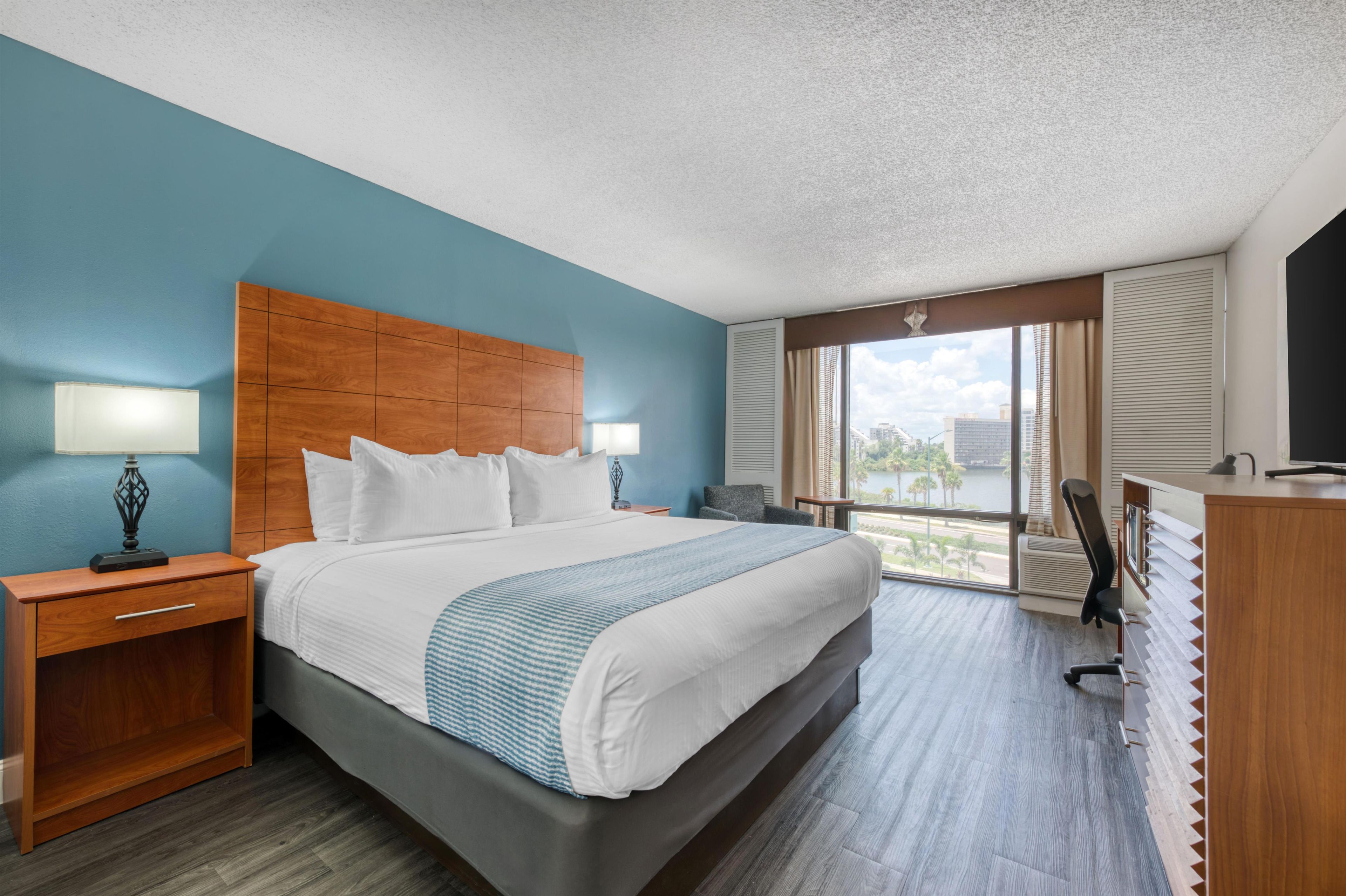 A hotel room with a large bed, a desk, a chair, a TV, and a window with a view of the city.