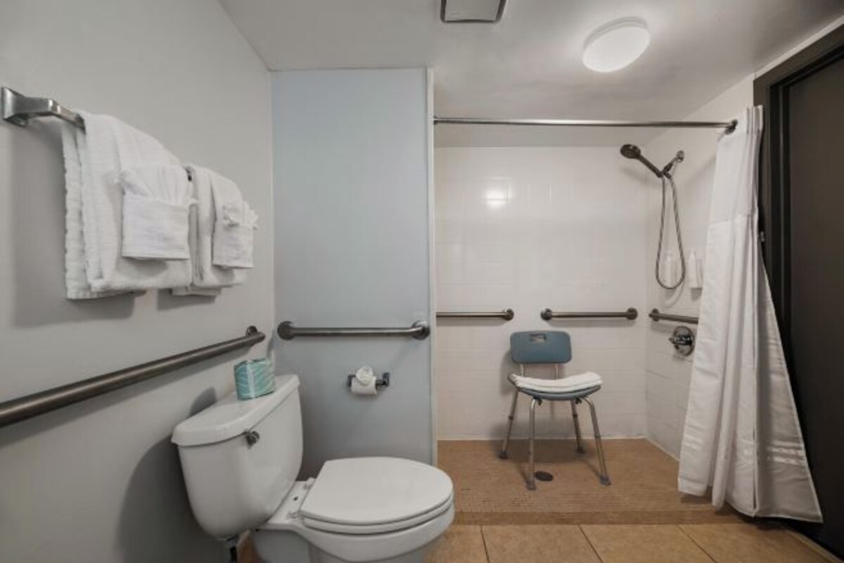 A bathroom with a toilet, handrails, a shower area, and a chair.