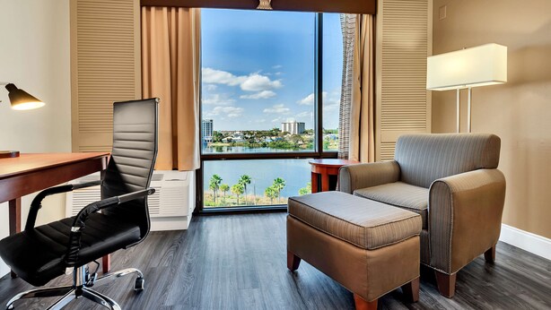 A hotel room with a large window, a desk, a black office chair, a brown sofa, and a lamp. - Best Western Orlando Gateway Hotel (Orlando)