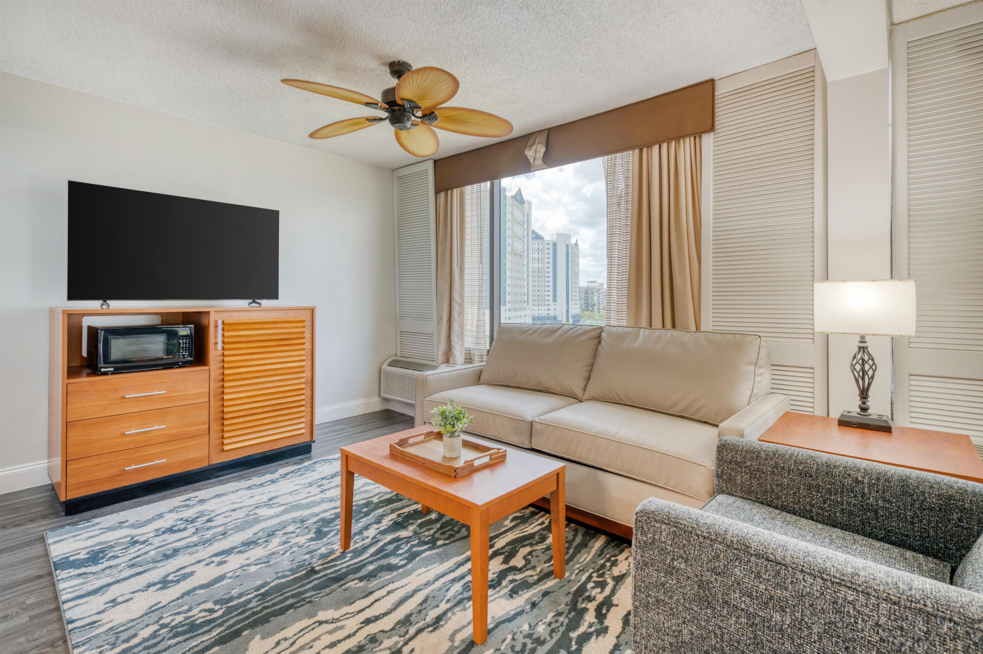 A modern hotel room with a flat-screen TV, a wooden cabinet, a sofa, a patterned rug, and a ceiling fan.