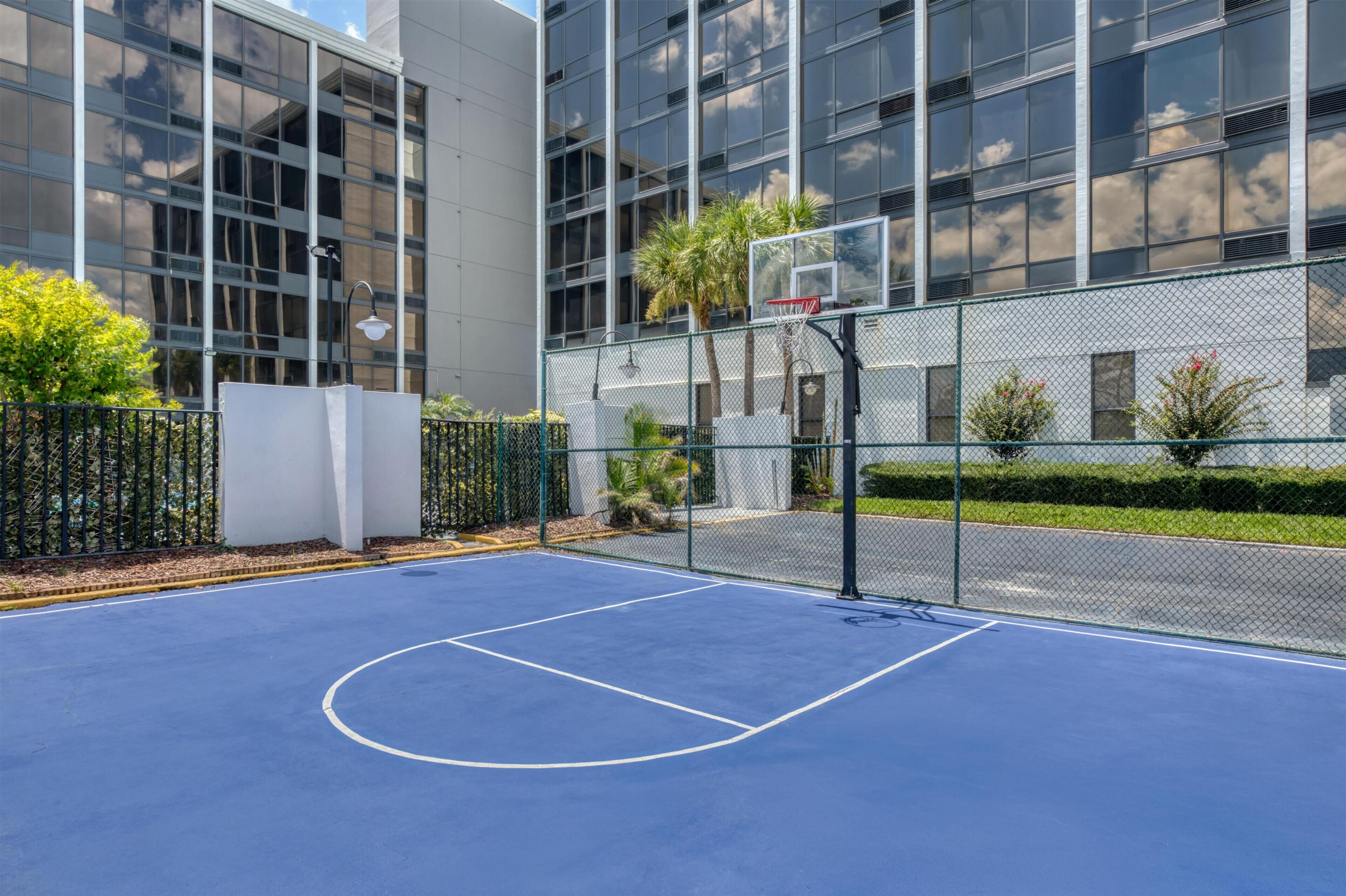 Un terrain de basket en plein air, entouré d’une clôture en treillis métallique, avec un panier de basket et un bâtiment aux grandes fenêtres en arrière-plan.