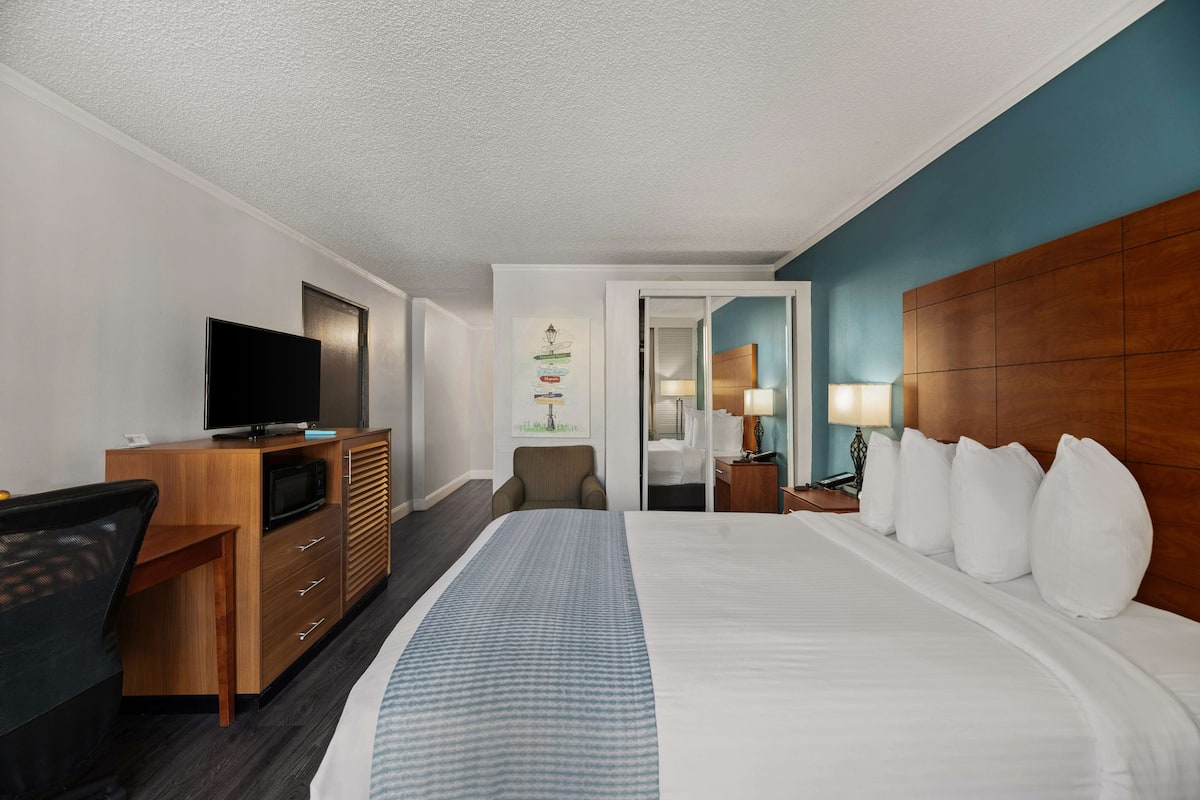 Een hotelkamer met een bed, een bureau met een televisie, een stoel, een nachtkastje met een lamp en een deur naar een andere kamer.