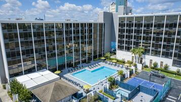 Un edificio moderno con piscina en la azotea, sillas de descanso y cancha de tenis.