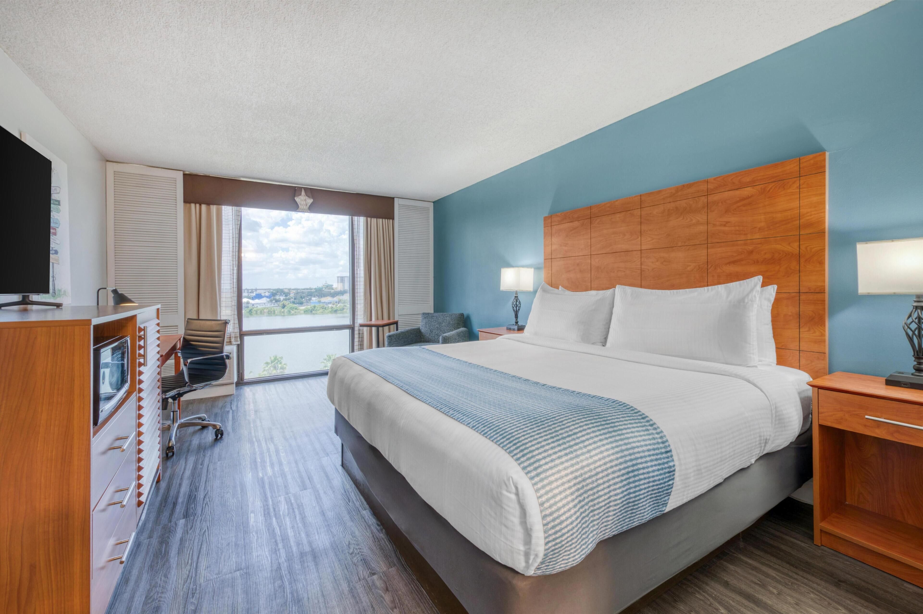 Habitación de hotel con una cama grande, un escritorio con computadora, una silla, televisor y vistas a la ciudad.