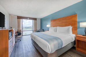 Hotellihuone, jossa on suuri sänky, työpöytä tietokoneella, tuoli, televisio ja näkymä kaupunkiin.