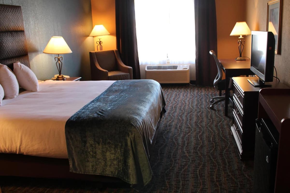 A hotel room with a bed, two bedside lamps, a chair, a desk with a TV, and an air conditioning unit.