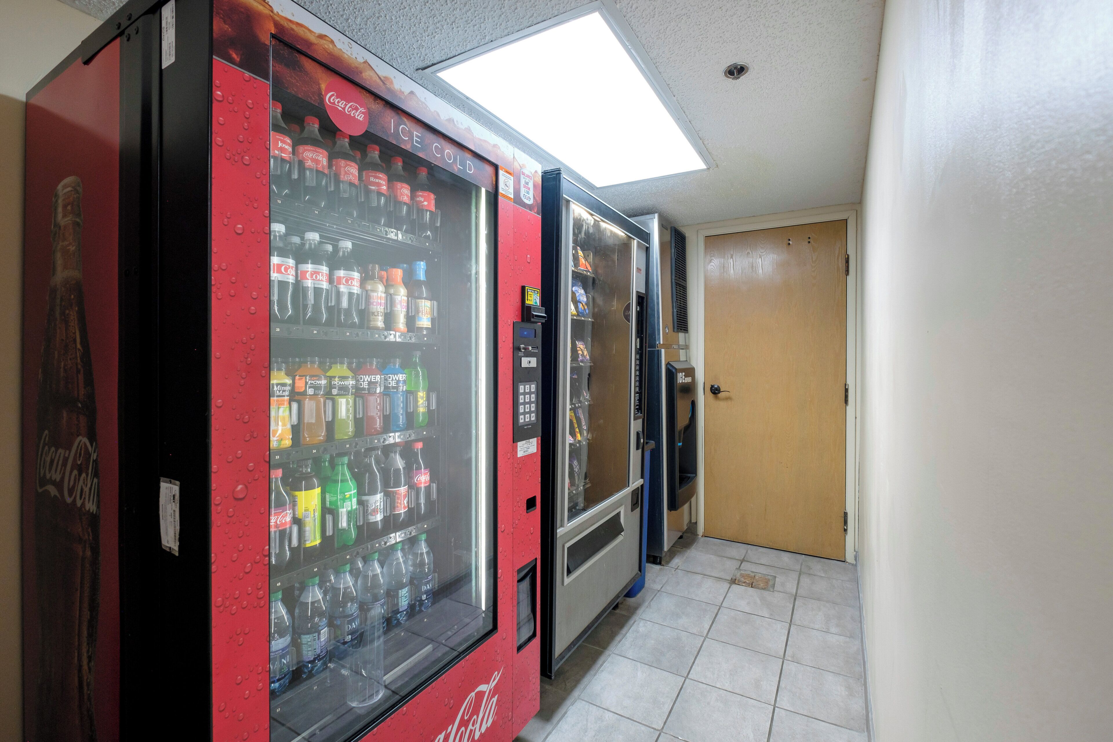 A vending machine area with multiple beverage dispensers, including Coca-Cola and other brands, in a hallway with tiled flooring.