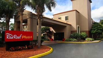 Entrada de un hotel Red Roof Plus con palmeras, un corredor cubierto y un área ajardinada con acolchado y plantas.