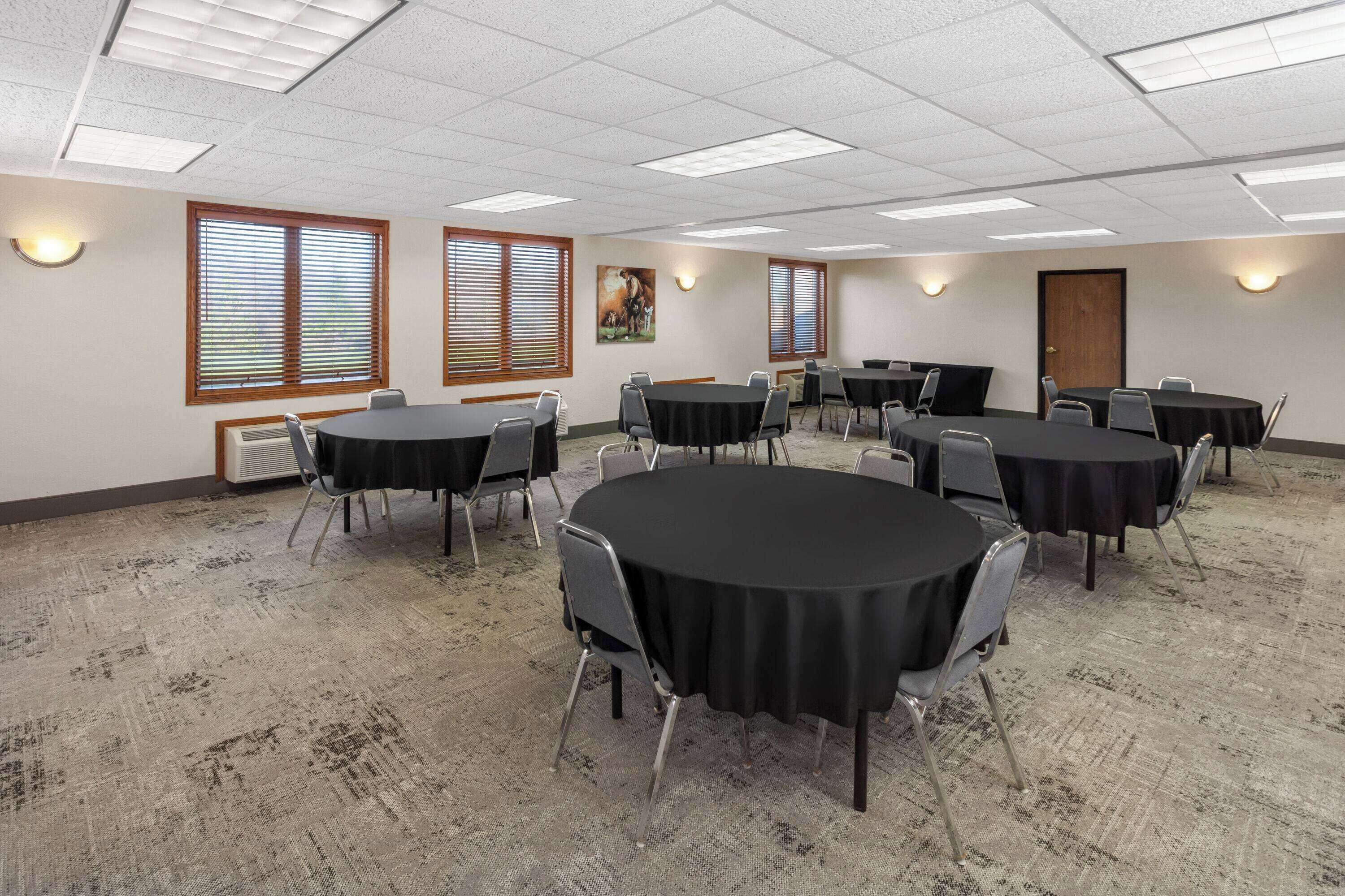 A conference room with multiple tables and chairs, large windows with blinds, and ceiling lights.