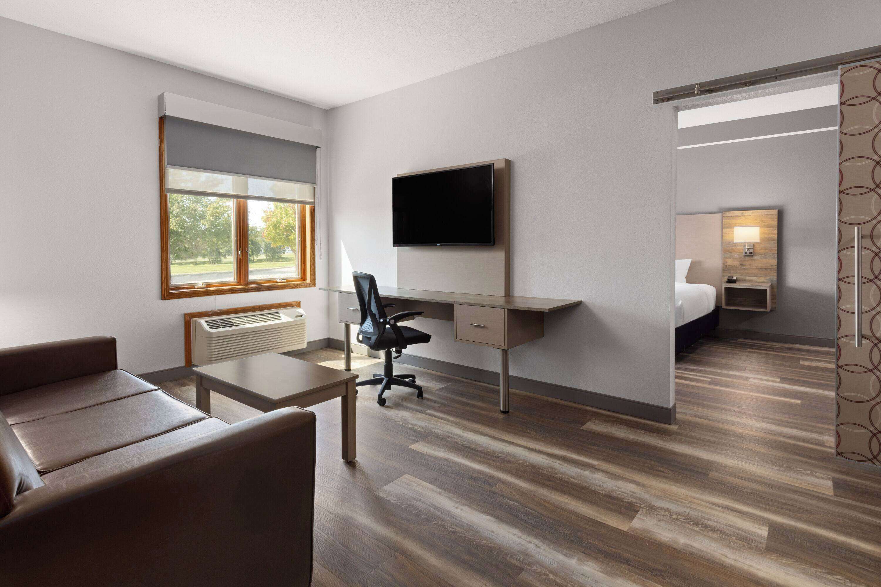 A modern hotel room with a desk, chair, sofa, TV, and a separate bedroom area.