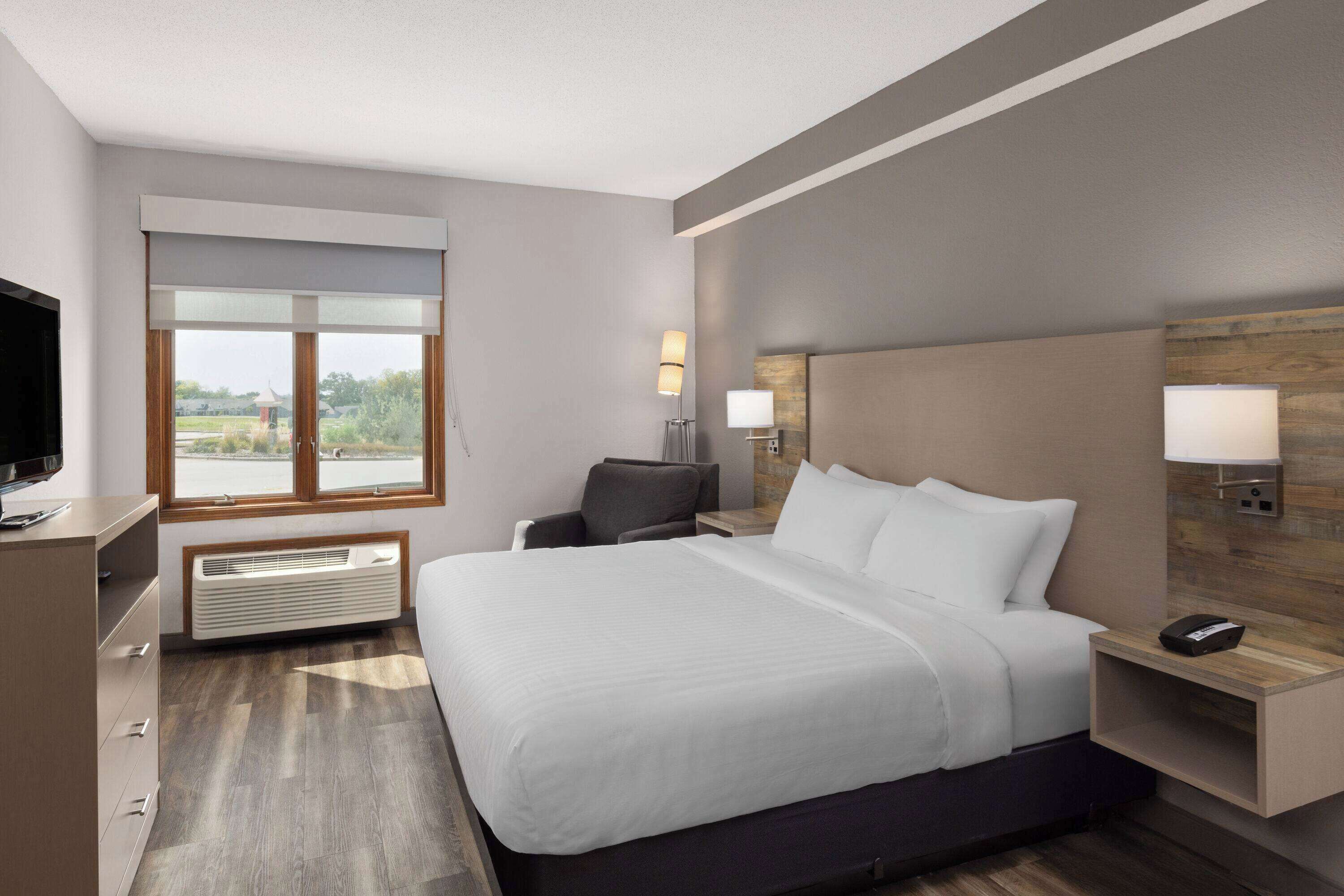 A modern hotel room with a large bed, a flat-screen TV, a desk, and a window with a view of greenery.