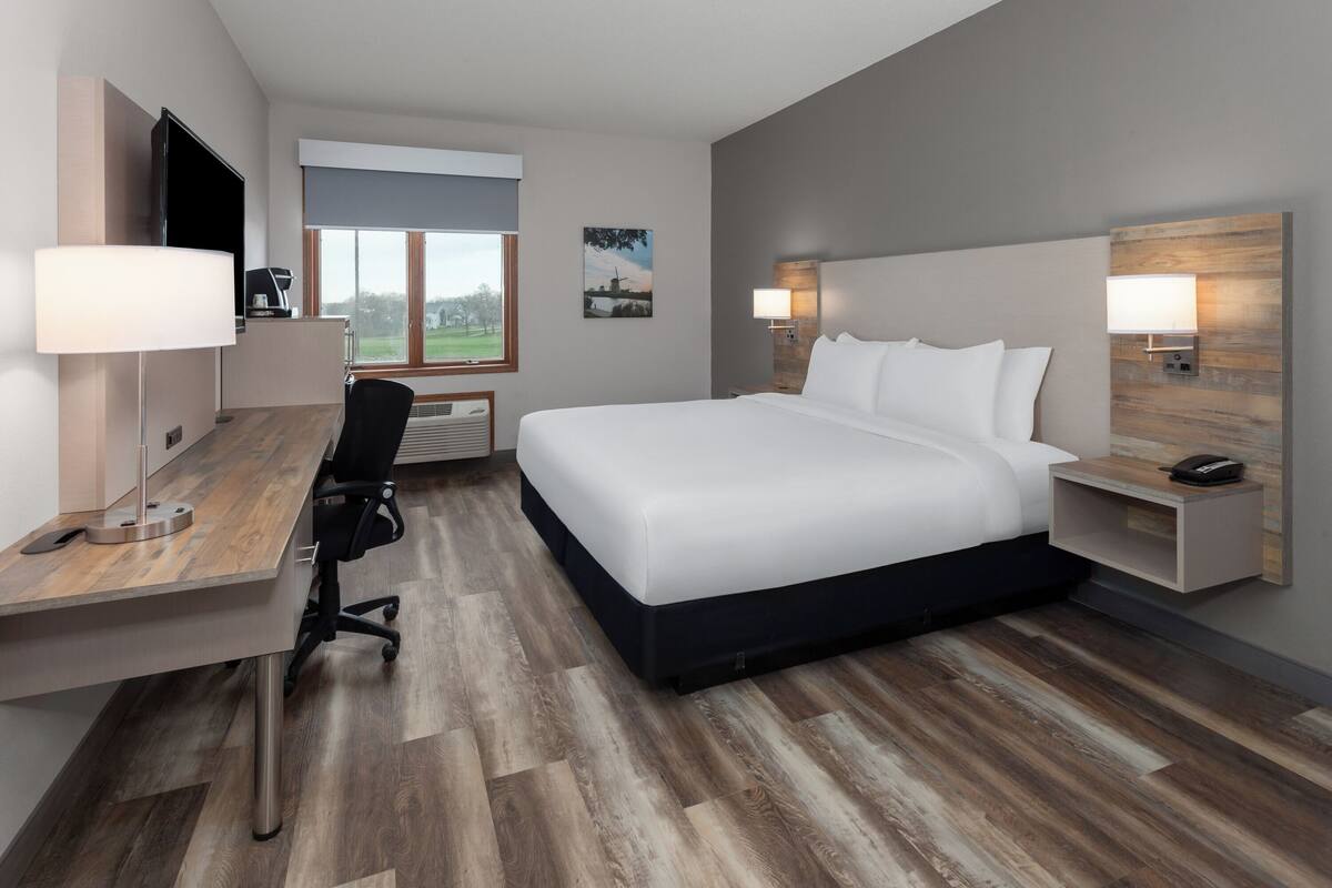 A modern hotel room with a large bed, a desk with a chair, a flat-screen TV, a coffee machine, and a window with a view of the outdoors.