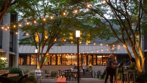 An outdoor seating area with string lights, tables, and chairs, overlooking a building with large windows.