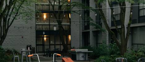 An outdoor seating area with a long wooden bench, tables, and chairs, surrounded by greenery and a building with large windows.