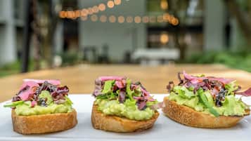 Three open-faced sandwiches with avocado and mixed greens on a white plate, set against a blurred background with string lights.