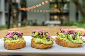 Trois sandwichs ouverts garnis d’avocat et d’un mélange de jeunes pousses, présentés sur une assiette blanche, le tout mis en valeur par un arrière-plan flou illuminé de guirlandes lumineuses.