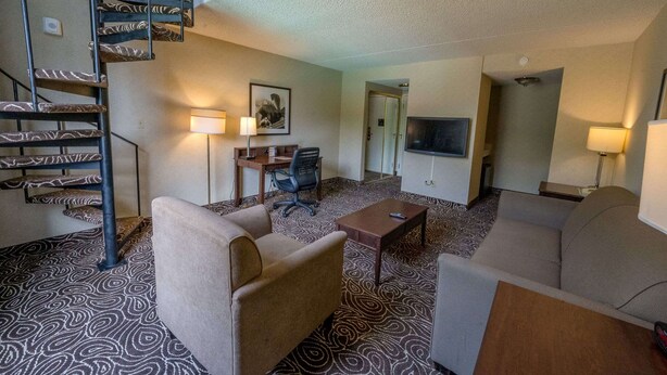 A hotel room with a patterned carpet, a sofa, a coffee table, a desk with a chair, a TV, and a staircase.