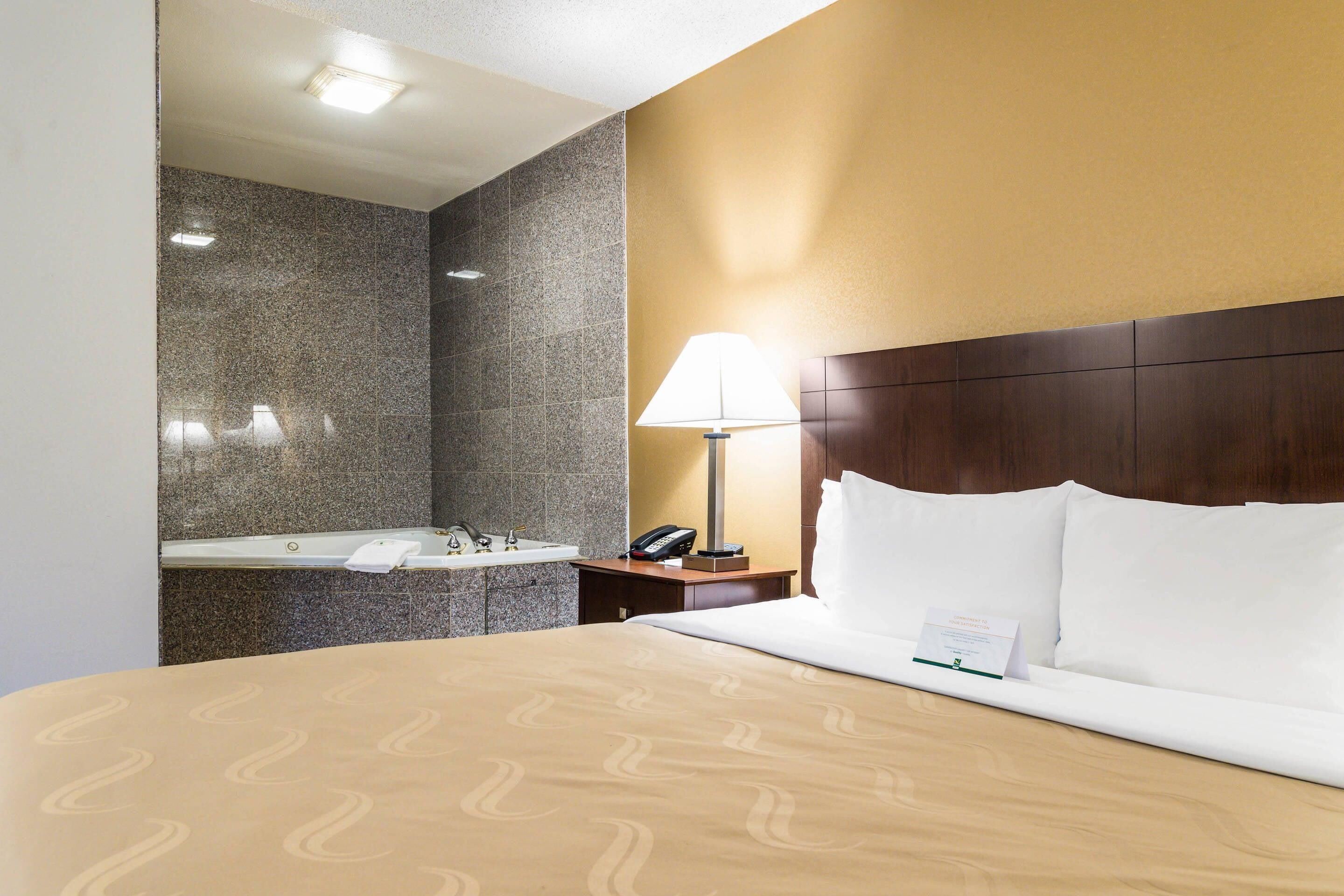 Quarto de hotel com cama, mesa de cabeceira, abajur, telefone e banheiro com pia e espelho.