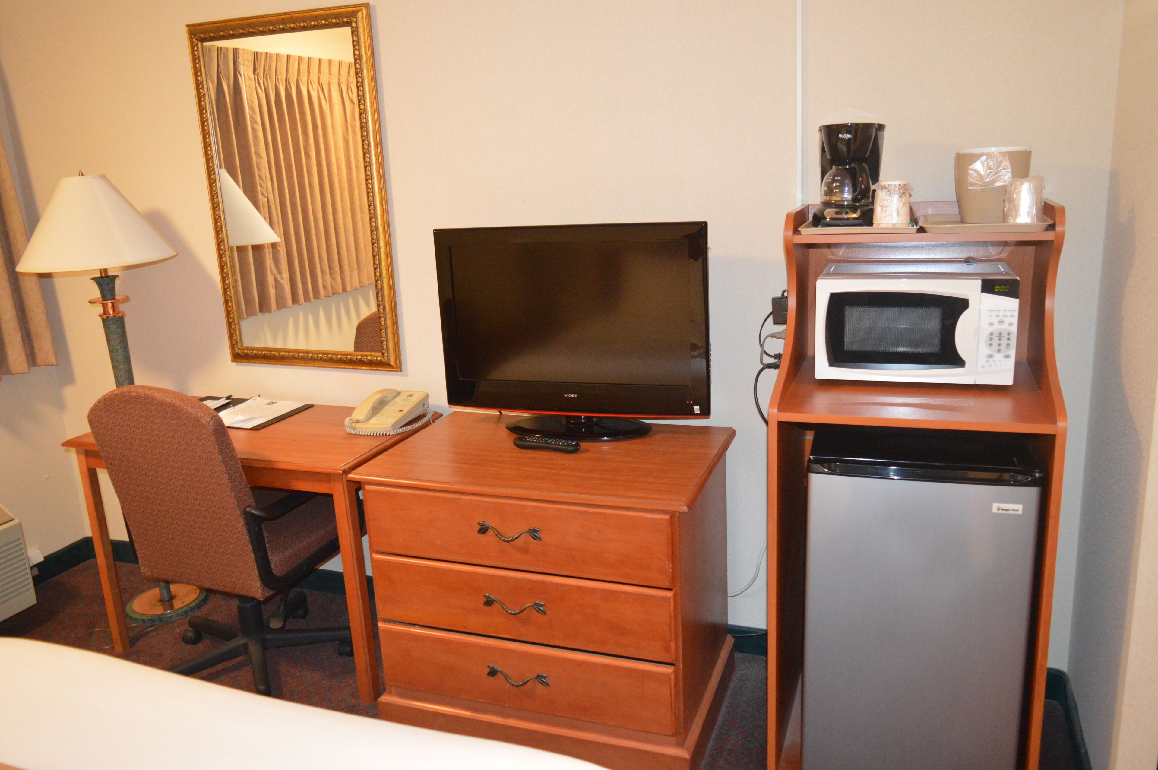 A hotel room with a wooden desk, a television, a microwave, a refrigerator, a chair, and a lamp.