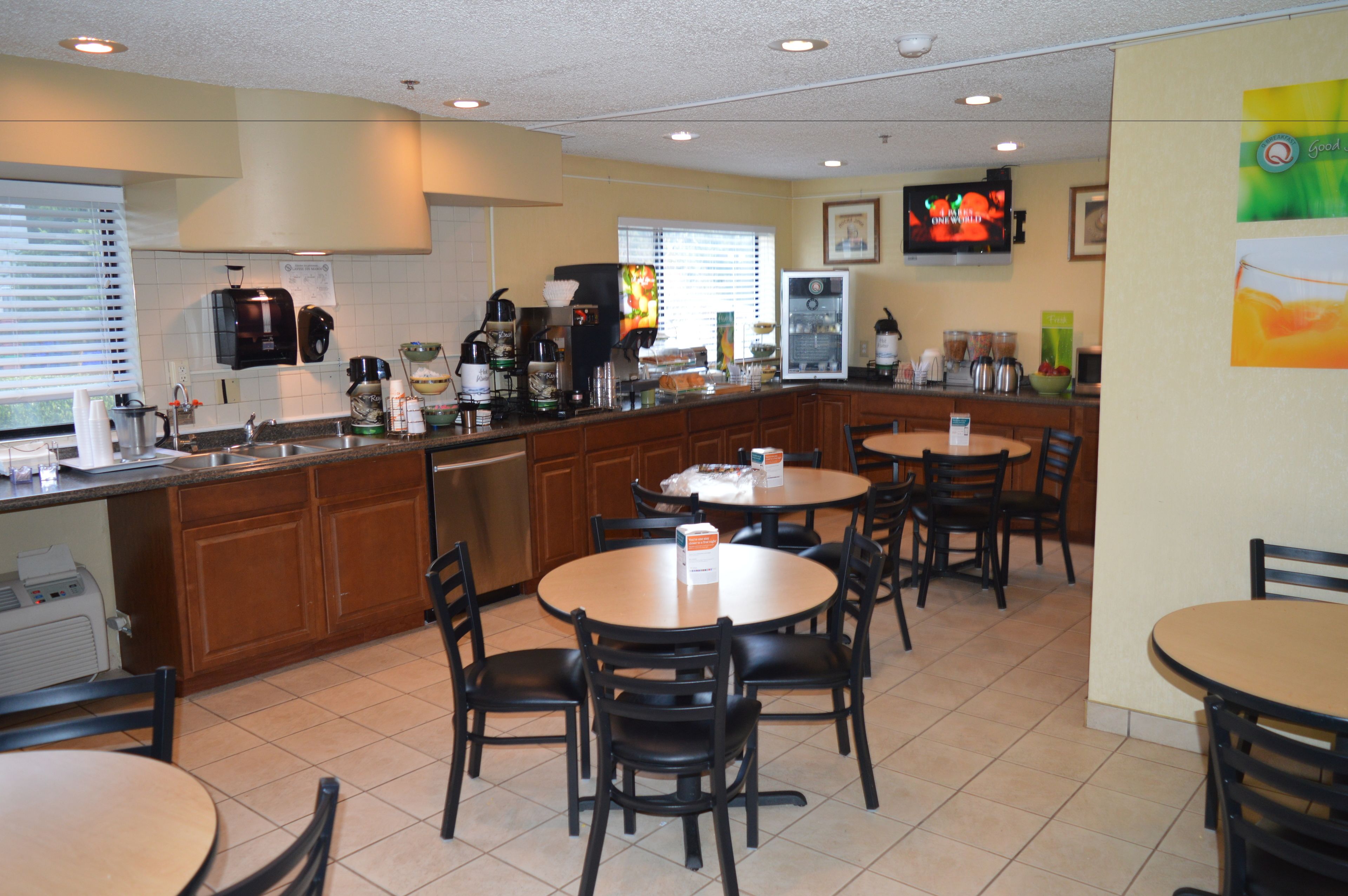 A restaurant with a kitchen area, dining tables, chairs, and a television mounted on the wall.