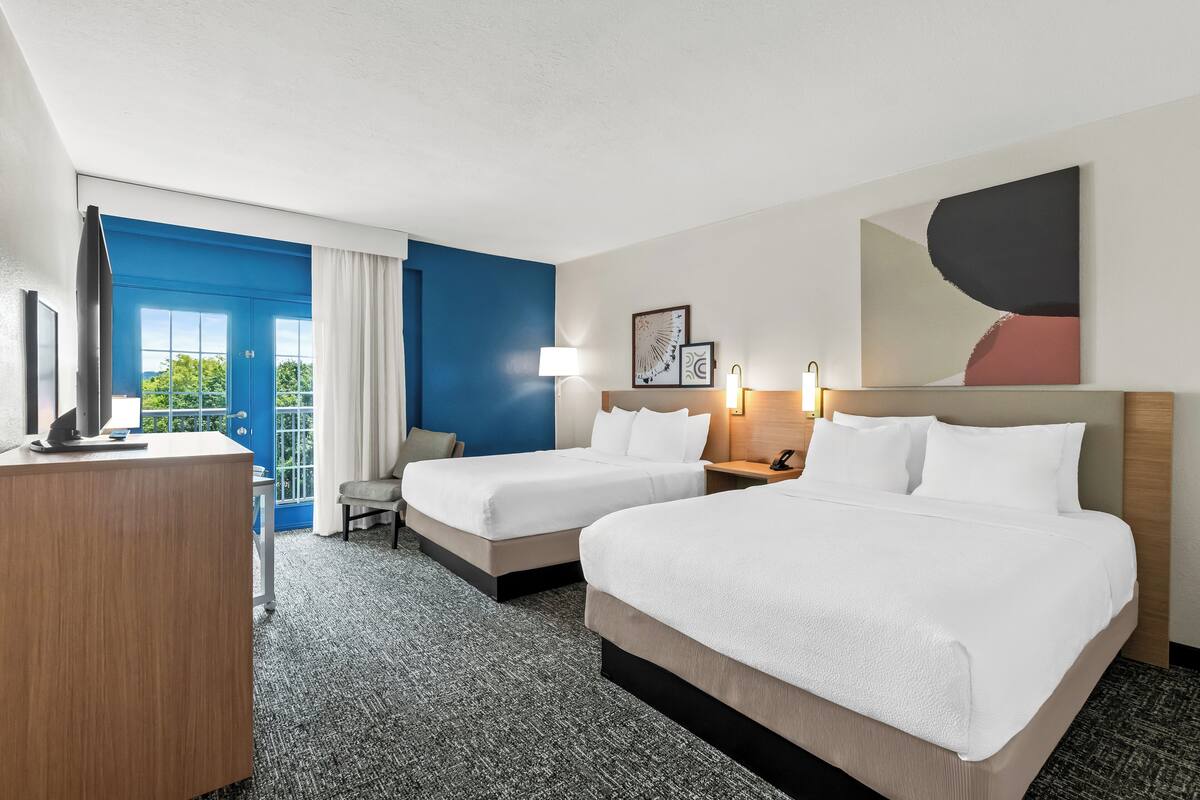 Ein Hotelzimmer mit zwei Betten, einem Schreibtisch, einem Sessel, einem Fernseher und einem großen Fenster mit Blick auf Grünflächen.