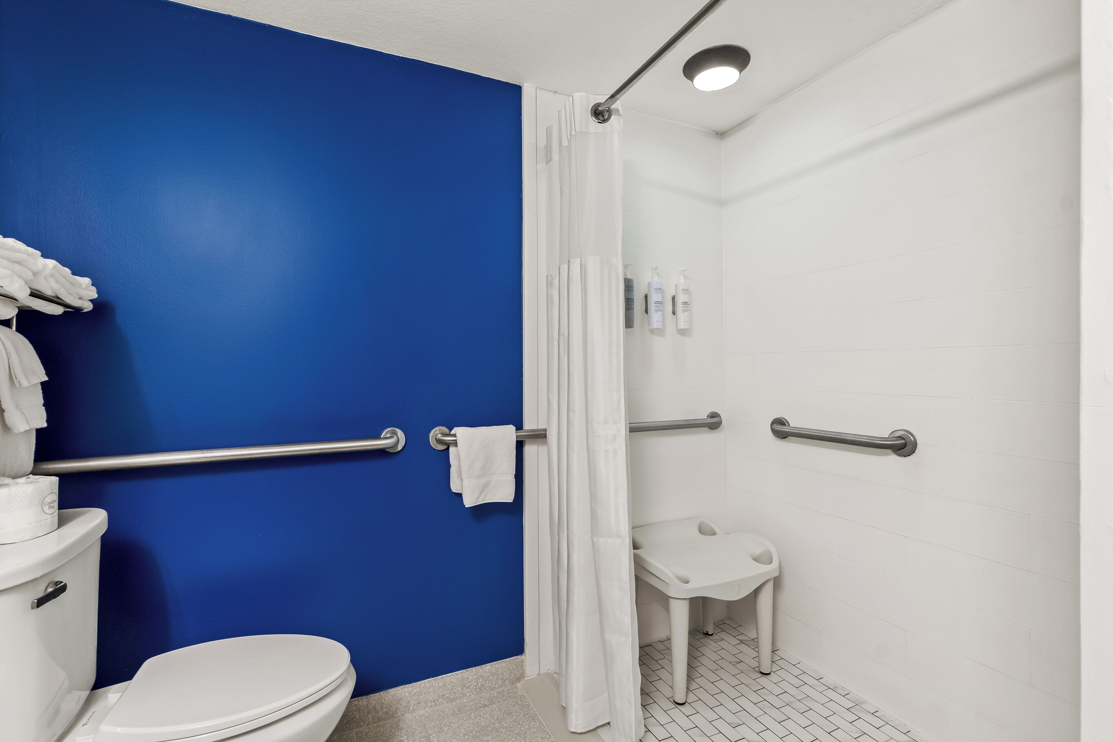 Un baño con una pared azul decorativa, un inodoro blanco, una ducha con cortina blanca y una barra de apoyo.