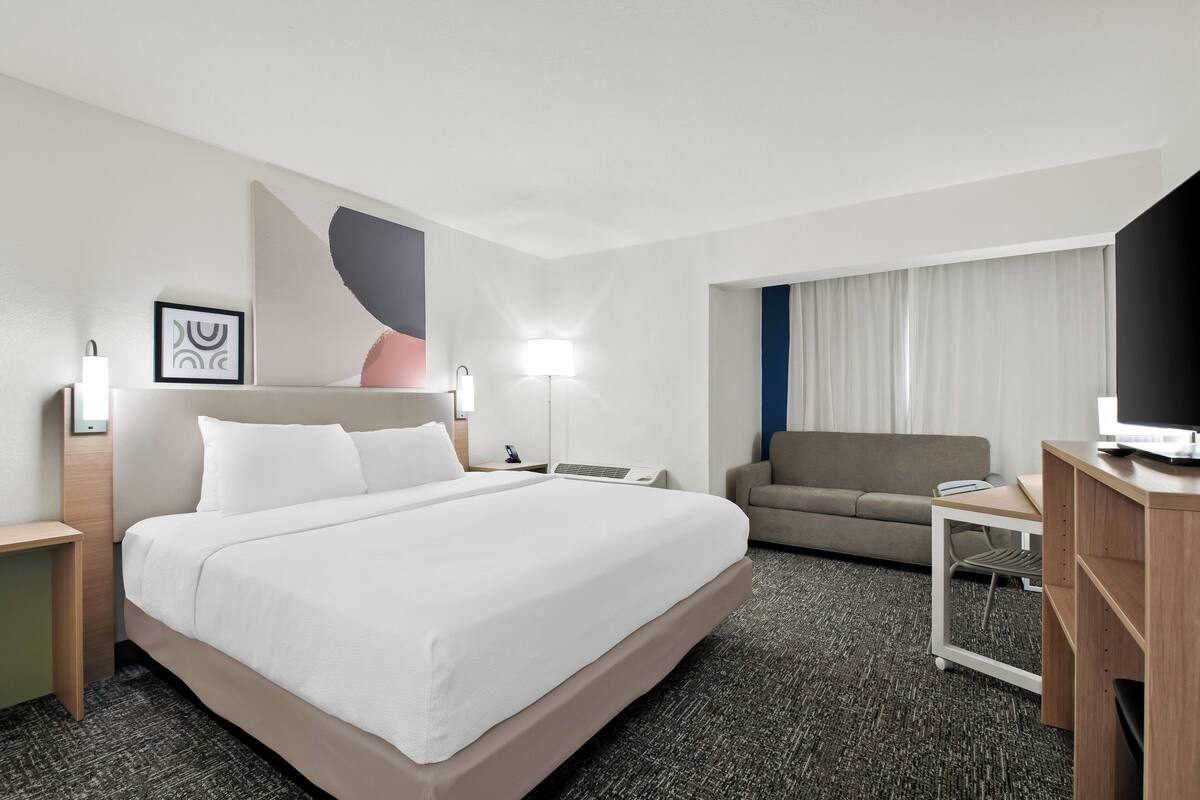 Ein Hotelzimmer mit einem großen Bett, einem Schreibtisch, einem Stuhl, einem Fernseher und einem Fenster mit Vorhängen.