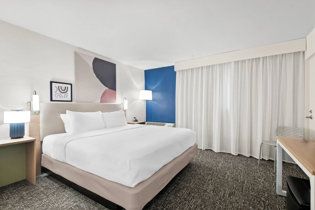 Ein Hotelzimmer mit einem großen Bett, einem Schreibtisch, einem Stuhl, einer Lampe, einem Bilderrahmen und einem Vorhang.