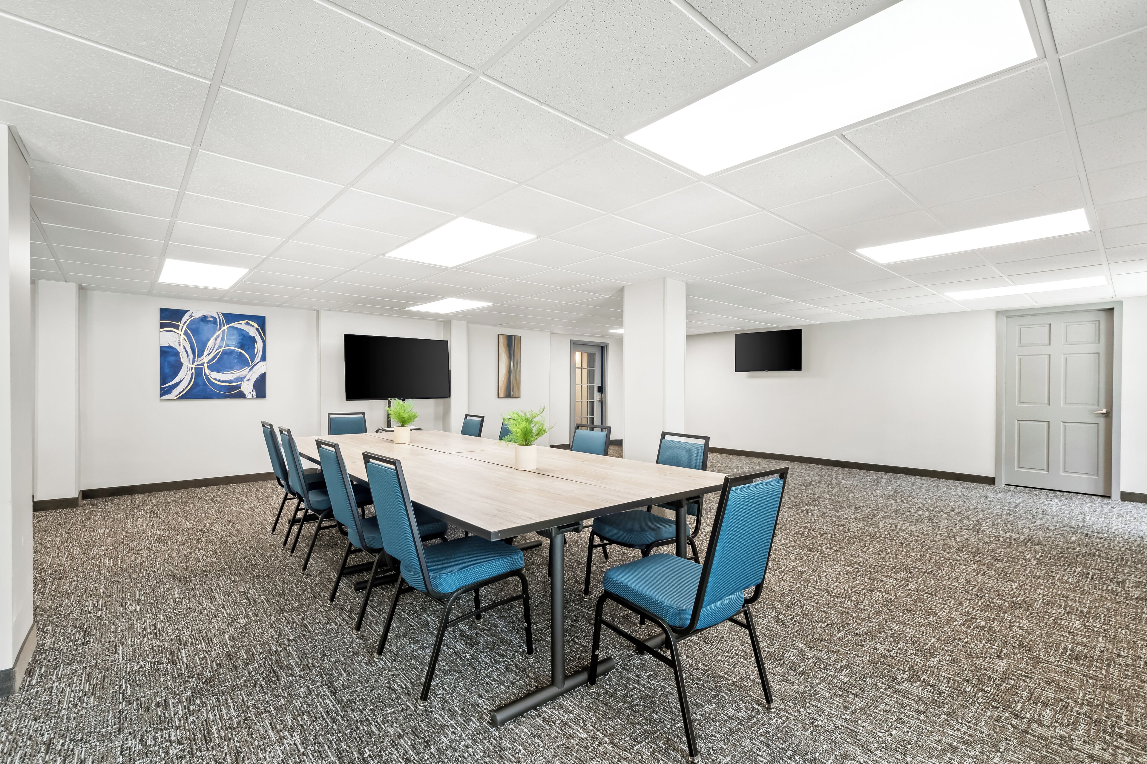 Un salón de conferencias con una larga mesa, sillas azules, un televisor de pantalla plana y un cuadro abstracto grande en la pared.