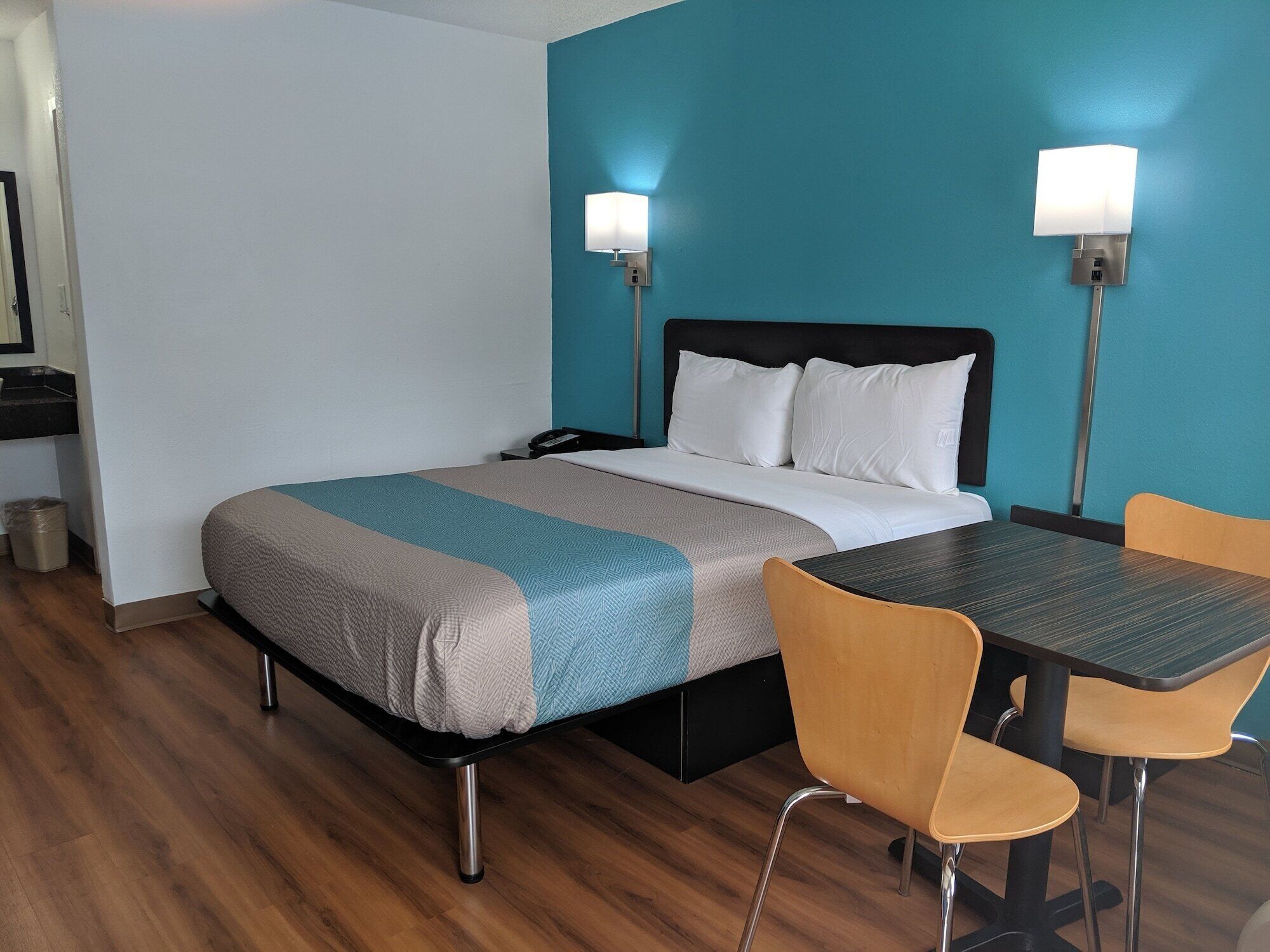 A hotel room with a bed, two bedside lamps, a desk with a chair, and a trash bin.