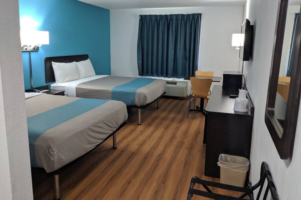 A hotel room with two beds, a desk with a computer, a chair, a desk lamp, a window with curtains, and a wall-mounted TV.