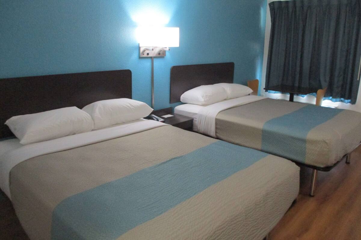 A hotel room with two beds, a nightstand, a lamp, and a window with curtains.