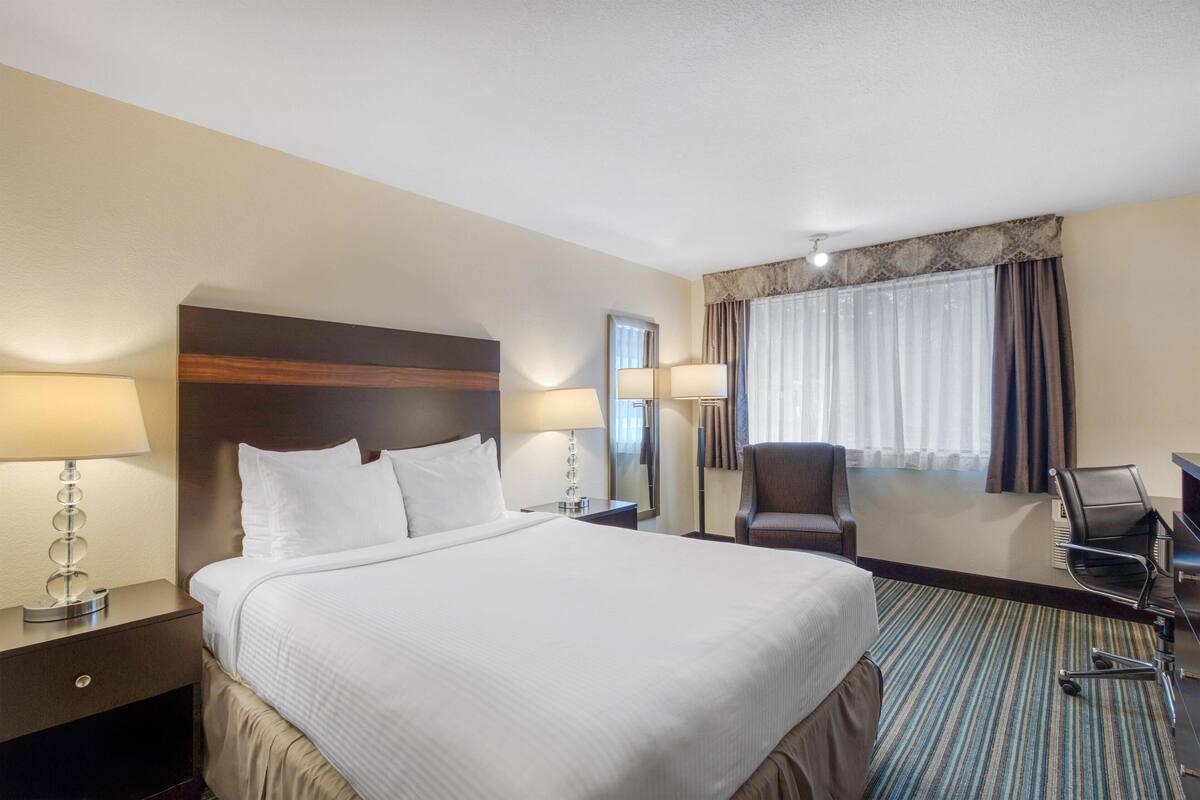 A hotel room with a large bed, two bedside tables with lamps, a desk with a chair, a large mirror, and a window with curtains.