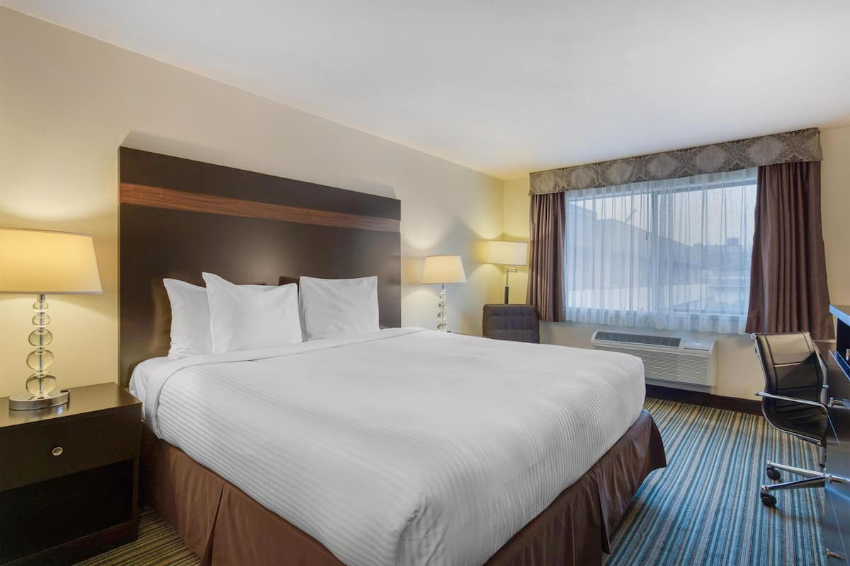 A hotel room with a large bed, two bedside tables with lamps, a desk with a chair, and a large window with curtains.